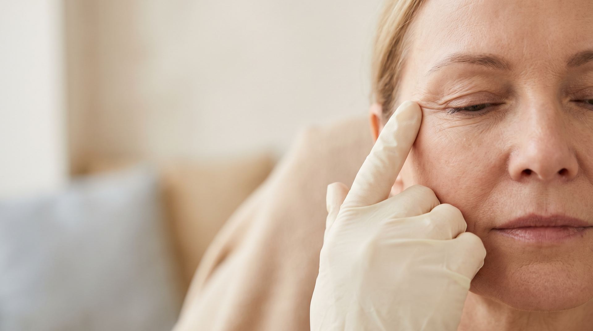 A clinician's gloved fingertip at the fine lines beside a woman's eye