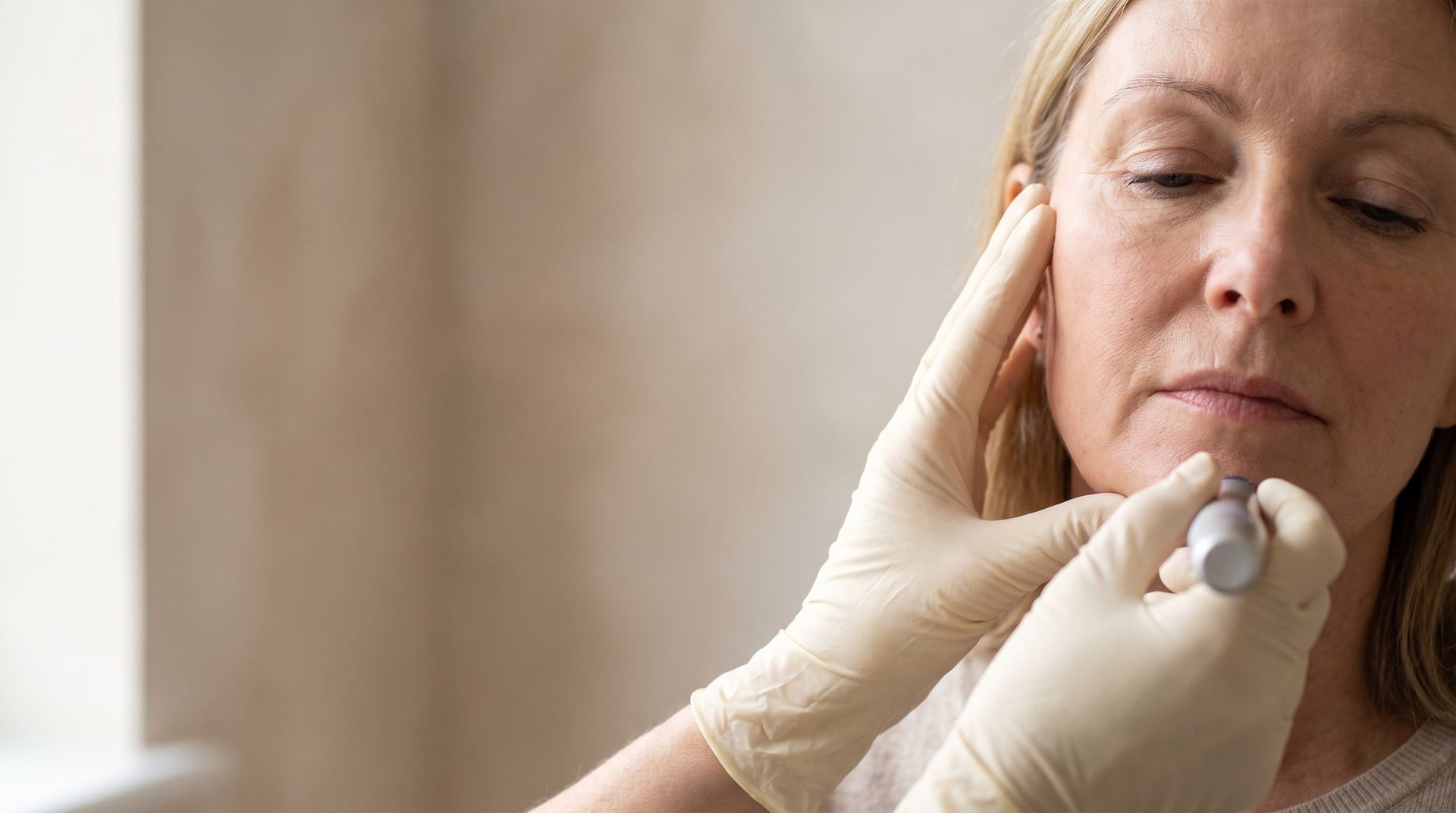 A clinician's gloved hands examining the jawline of a woman in her fifties
