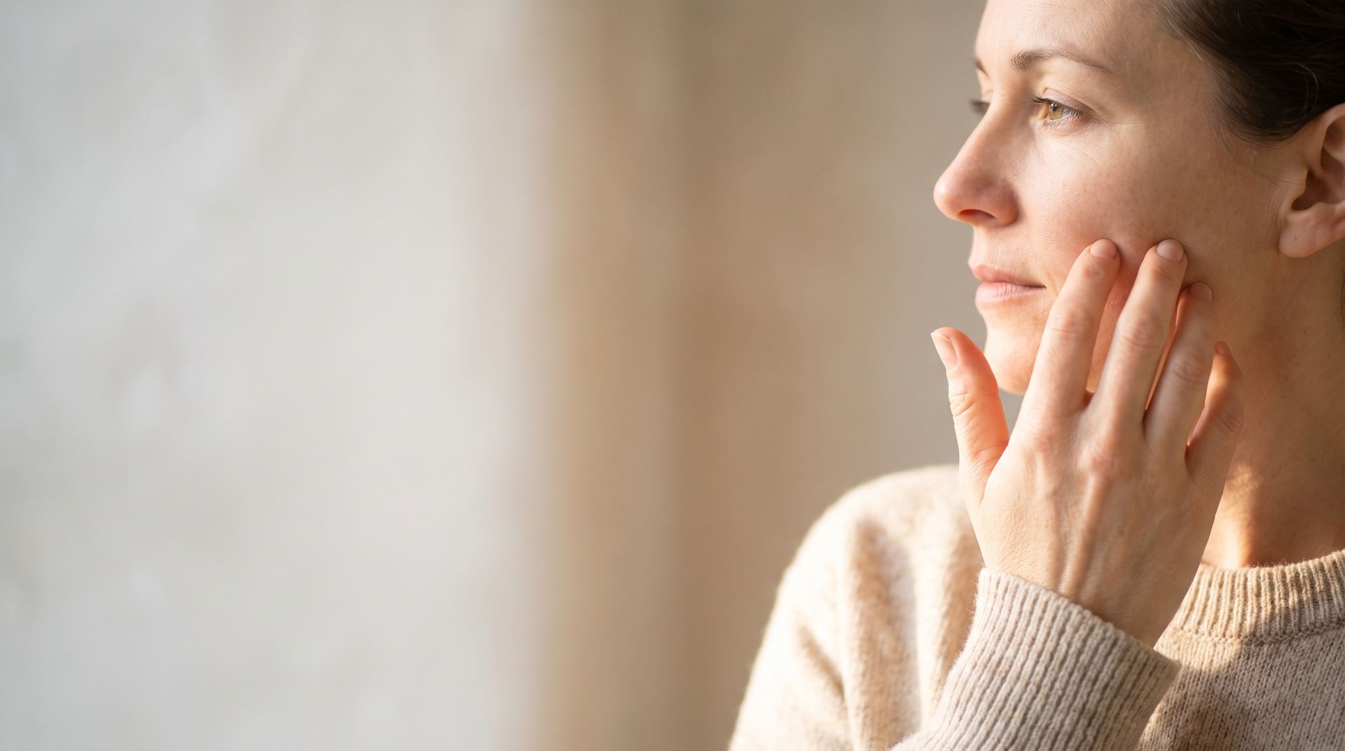 A woman in a cream jumper lightly touching her cheek