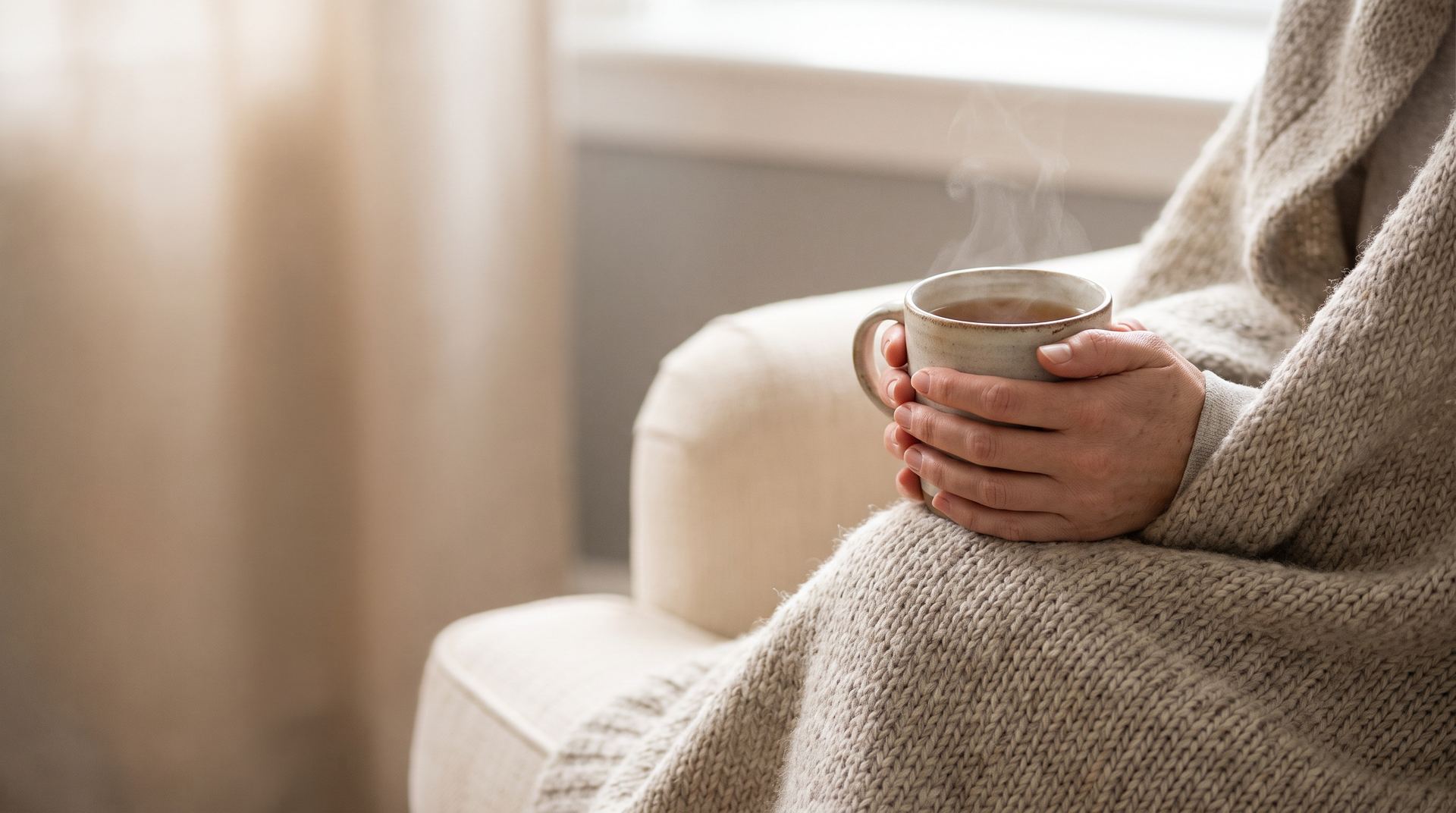 Hands wrapped around a cup of tea, a knitted blanket over the knees