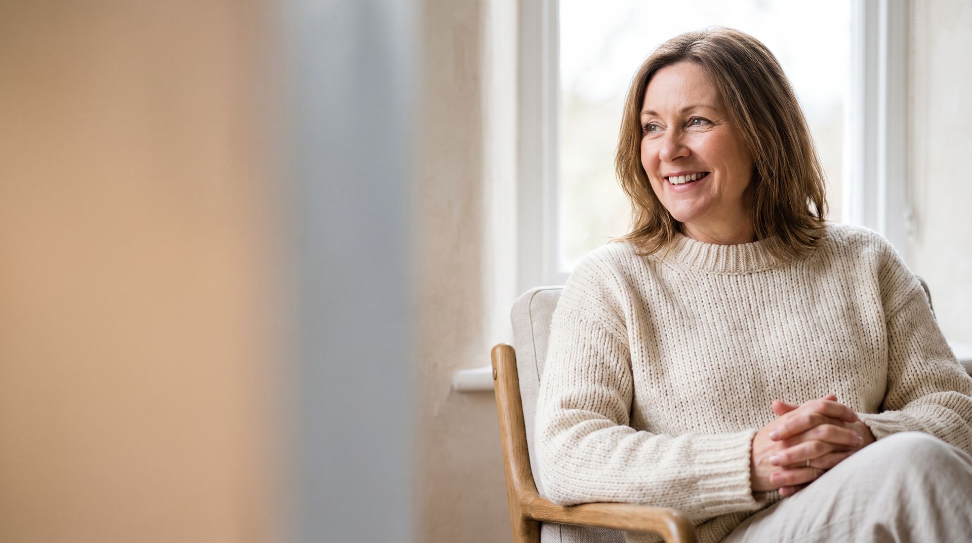 A smiling woman in a cream jumper by a window