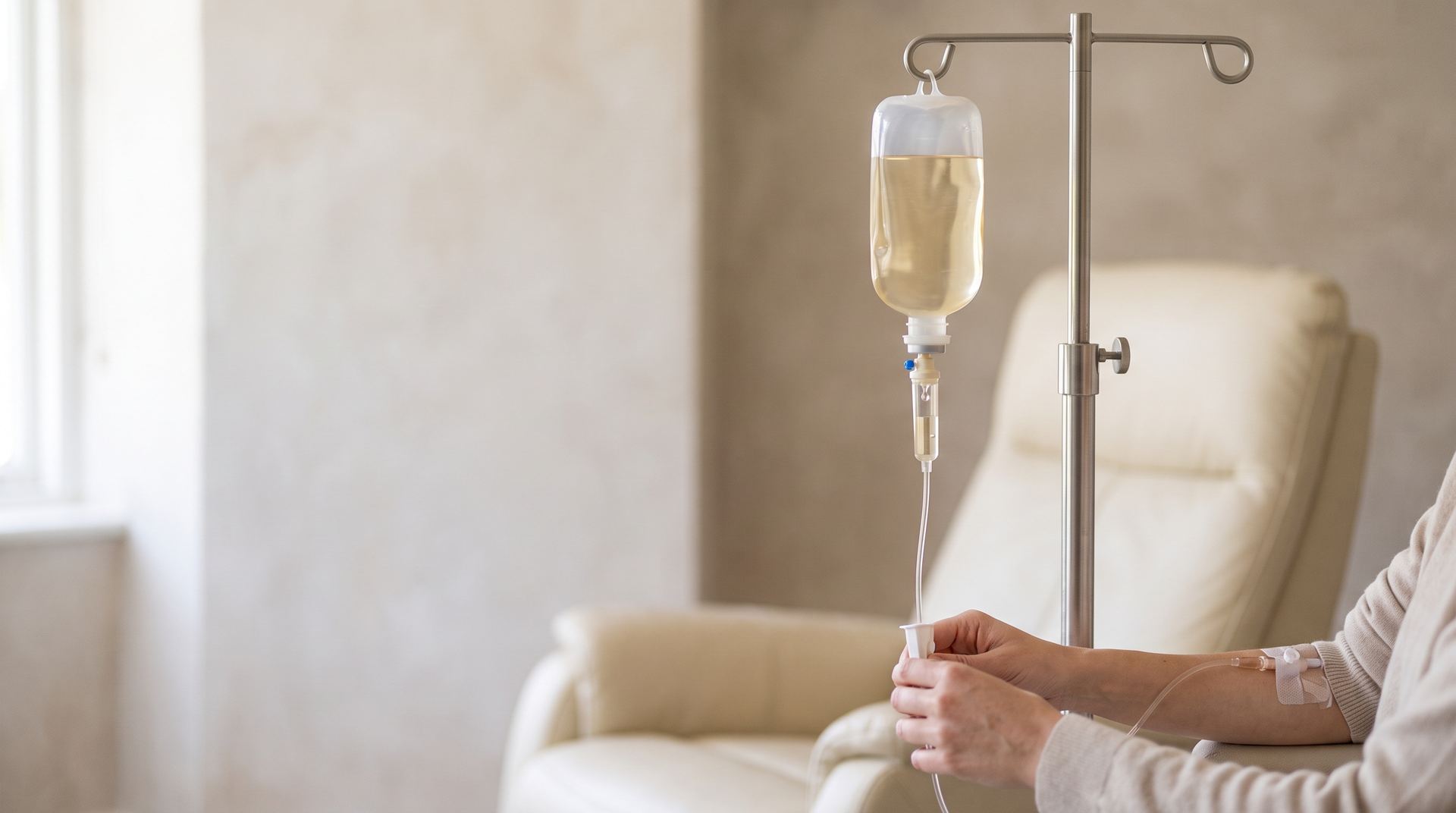 An infusion of pale golden vitamin fluid on a drip stand beside a patient resting in a cream recliner