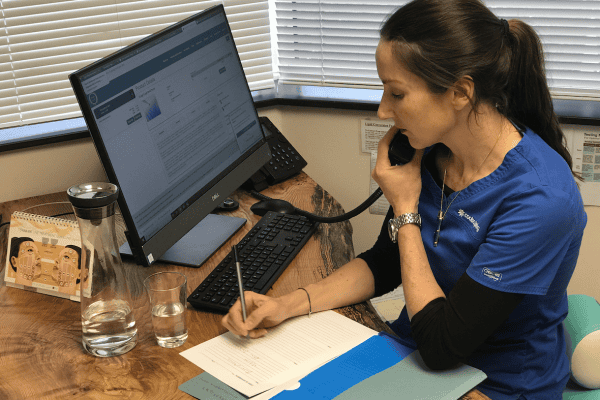 Annelize Meyer in a blue uniform is speaking on the phone while taking notes at a desk with a computer and a glass of water, ensuring online consultations provide a lifeline to new and existing patients at Meyer Clinic. - Meyer Clinic