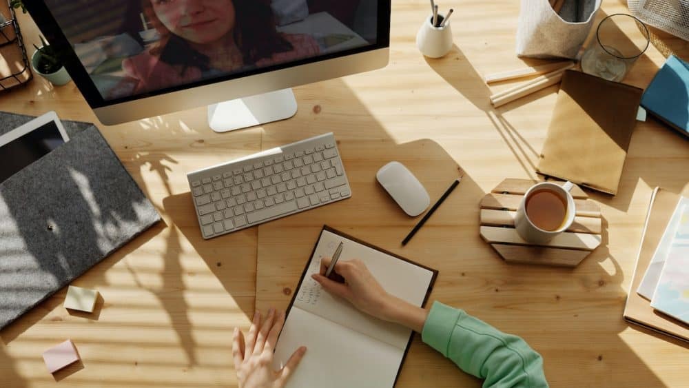 Person taking notes at a wooden desk during a video call on a computer monitor, jotting down tips on staying fit and healthy while working from home. Desk items include a notebook, keyboard, mouse, coffee cup, and folders. - Meyer Clinic
