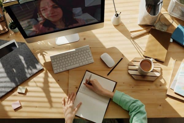 Person taking notes at a wooden desk during a video call on a computer monitor, jotting down tips on staying fit and healthy while working from home. Desk items include a notebook, keyboard, mouse, coffee cup, and folders. - Meyer Clinic