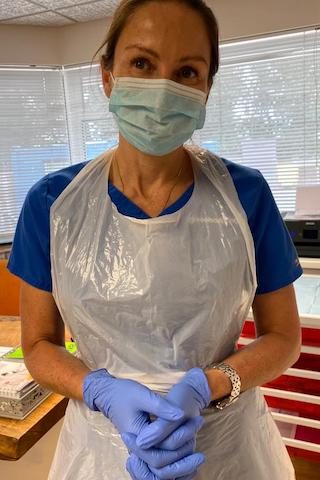 Annelize Meyer wearing a mask, gloves, and apron stands in a brightly lit room with blinds. She is facing the camera with hands clasped. - Meyer Clinic
