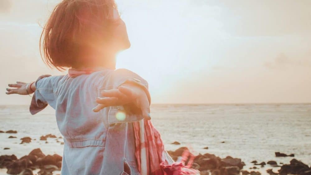 Person with outstretched arms facing the sun at a rocky beach during sunset, embracing nature’s serene beauty—a perfect moment to boost your oestrogen levels naturally. - Meyer Clinic