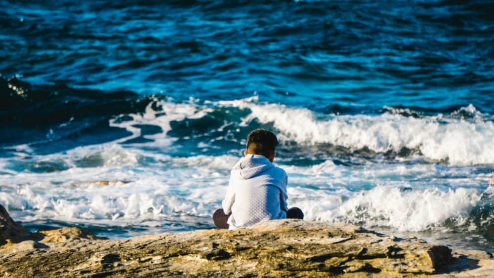 A person in a hoodie sits on a rocky shoreline, embracing mindfulness as the oceans waves crash—a serene escape reminiscent of teaching mindfulness to children during lockdown. - Meyer Clinic