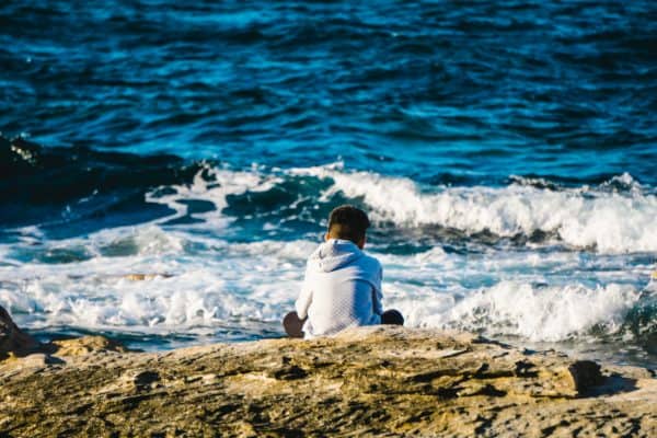 A person in a hoodie sits on a rocky shoreline, embracing mindfulness as the oceans waves crash—a serene escape reminiscent of teaching mindfulness to children during lockdown. - Meyer Clinic