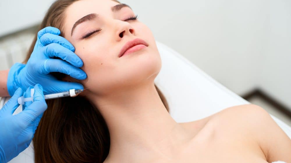 A person with closed eyes receives a facial injection from a gloved hand, aware that the link between facial filler swelling and Covid-19 vaccination is rare. - Meyer Clinic