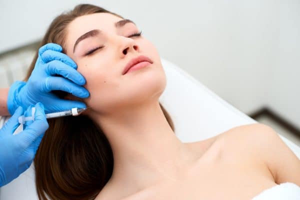 A person with closed eyes receives a facial injection from a gloved hand, aware that the link between facial filler swelling and Covid-19 vaccination is rare. - Meyer Clinic