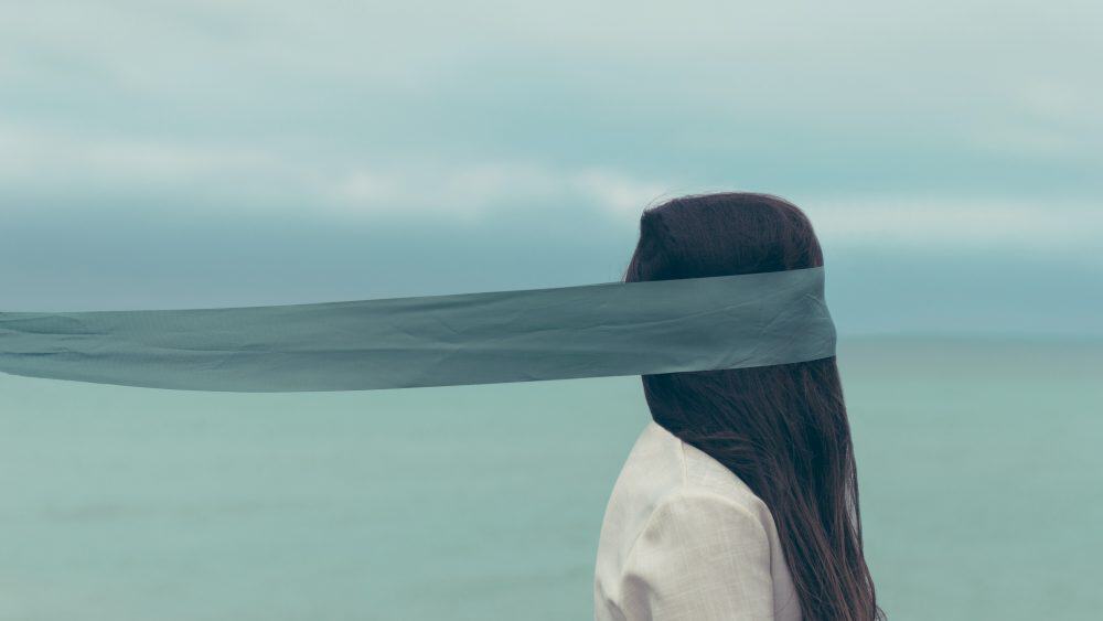 A teen with long hair stands by the ocean, partially obscured by a sheer fabric covering their eyes, embraced by the gentle complexity of being under a cloudy sky. - Meyer Clinic