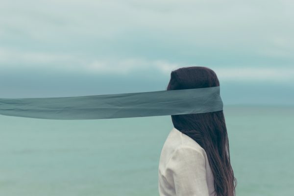 A teen with long hair stands by the ocean, partially obscured by a sheer fabric covering their eyes, embraced by the gentle complexity of being under a cloudy sky. - Meyer Clinic