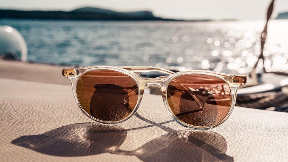 Clear-framed sunglasses with brown lenses rest on a boat seat, soaking in the warm sunlight while overlooking a sunny, sparkling body of water with blurred distant hills. - Meyer Clinic