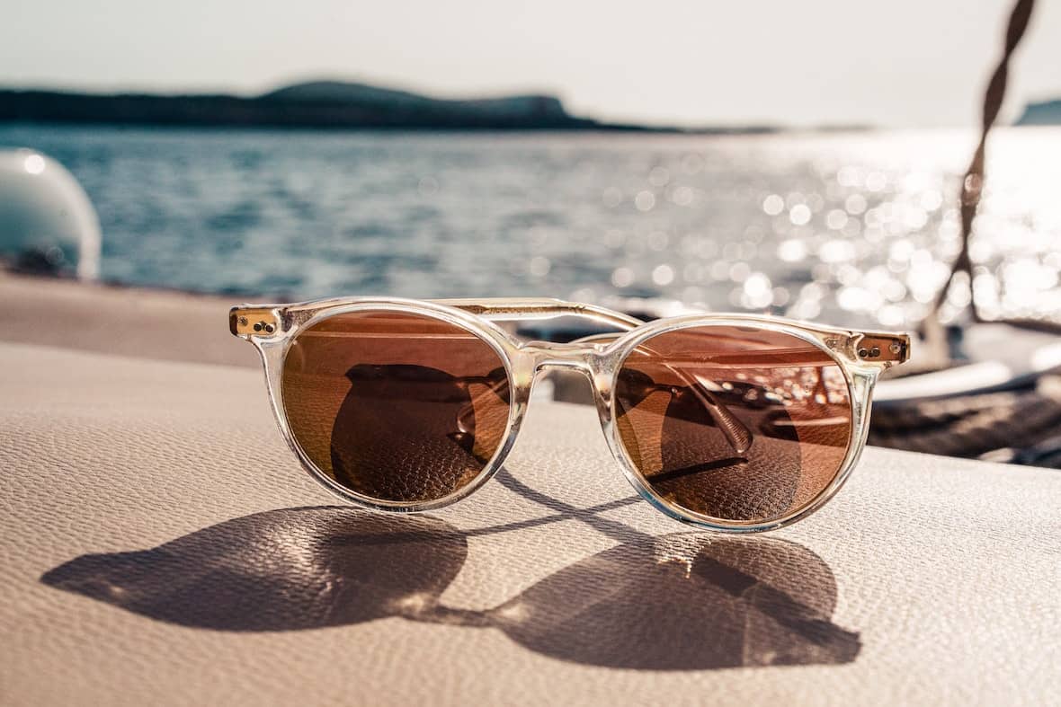 Clear-framed sunglasses with brown lenses rest on a boat seat, soaking in the warm sunlight while overlooking a sunny, sparkling body of water with blurred distant hills. - Meyer Clinic