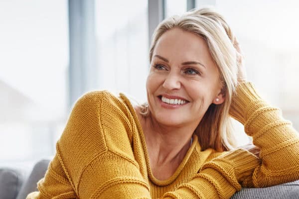 A person with long blonde hair, wearing a yellow sweater, smiles while leaning on a sofa, exuding warmth and calmness. A bright window in the background hints at rejuvenation and menopause help in this peaceful setting. - Meyer Clinic