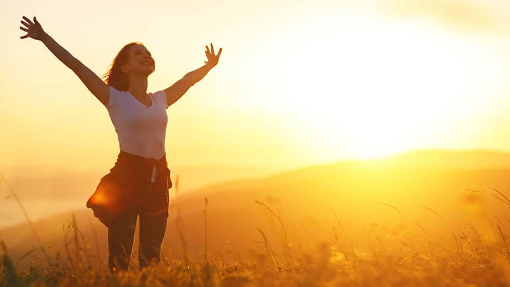 A person stands in a field with arms outstretched, facing a bright sunrise or sunset, contemplating the rejuvenating possibilities at Meyer Clinic. - Meyer Clinic