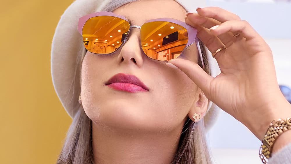 A woman wearing cat-eye sunglasses adjusts them while looking up. With pink lipstick and a light beret, her hair is partially visible, hinting at the glow one might achieve from a superficial chemical peel. - Meyer Clinic