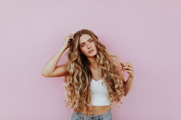 A woman with long, wavy hair poses confidently against a pink wall, showcasing her fresh look in a white top and jeans. Perhaps its the work of Ultherapy®, adding an extra glow to her effortless style. - Meyer Clinic
