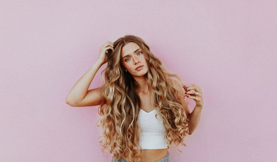 A woman with long, wavy hair poses confidently against a pink wall, showcasing her fresh look in a white top and jeans. Perhaps its the work of Ultherapy®, adding an extra glow to her effortless style. - Meyer Clinic