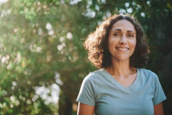 A woman with curly hair smiles at the camera, standing outdoors with sunlight filtering through trees in the background, her face glowing like a subject from Dr. Meyers recent research. - Meyer Clinic