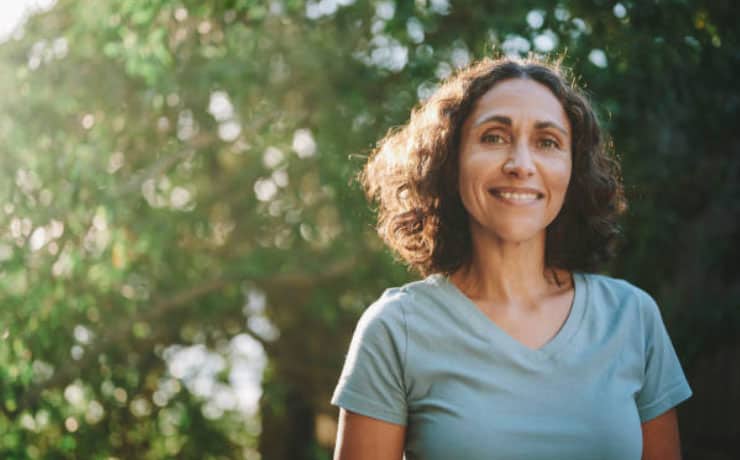 A woman with curly hair smiles at the camera, standing outdoors with sunlight filtering through trees in the background, her face glowing like a subject from Dr. Meyers recent research. - Meyer Clinic