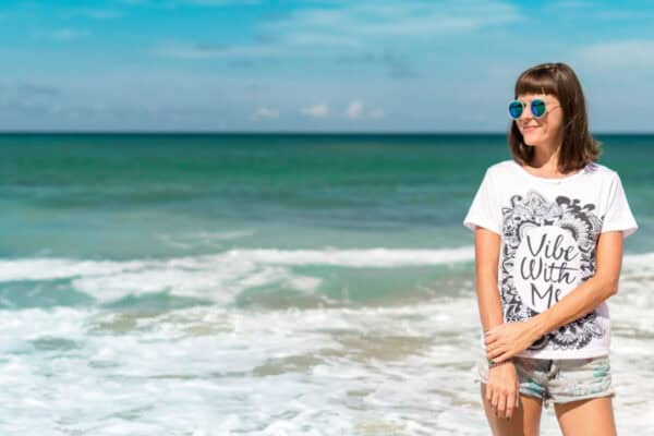 A woman stands on a beach, wearing sunglasses, a white T-shirt with a design about Hormone Health, and shorts. The ocean and sky provide a serene backdrop. - Meyer Clinic
