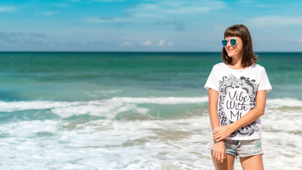 A woman stands on a beach, wearing sunglasses, a white T-shirt with a design about Hormone Health, and shorts. The ocean and sky provide a serene backdrop. - Meyer Clinic