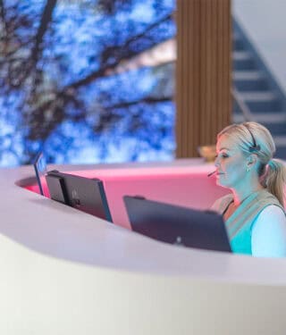 A woman with a headset sits at a curved desk with computer monitors, immersing herself in the patient journey against the backdrop of a large tree image. - Meyer Clinic